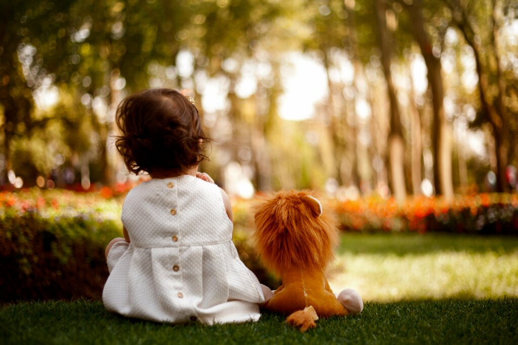 A child in a white dress sits on grass with a toy lion, enjoying a sunny day in the park.